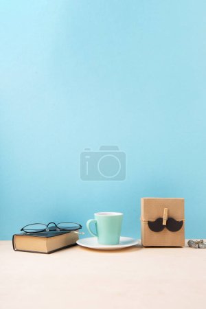 Téléchargez les photos : Fathers day holiday card concept with craft gift box, moustache noire, tasse de café, livre et verres sur fond bleu, espace de copie. - en image libre de droit