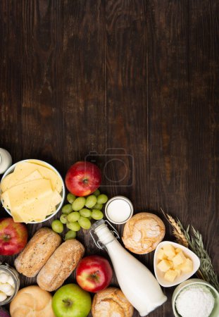 Happy Shavuot festive card. Jewish religious holiday concept. Dairy products, fruits, cheese, bread, milk bottle, cottage cheese, wheat on dark wooden background. Top view, copy space.