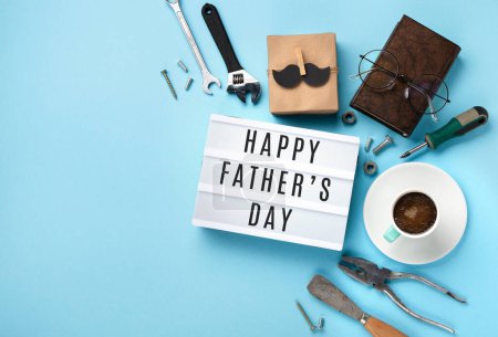 Photo for Fathers day holiday card concept with craft gift box, black moustache, cup of coffee, tools, book, empty white lightbox and glasses on blue background, top view, copy space. - Royalty Free Image