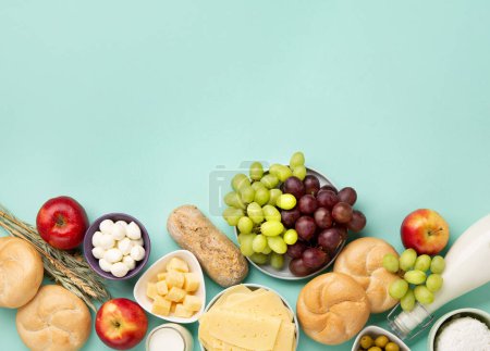 Happy Shavuot festive card. Jewish religious holiday concept. Dairy products, fruits, cheese, bread, milk bottle, cottage cheese, wheat on blue mint background. Top view, copy space.