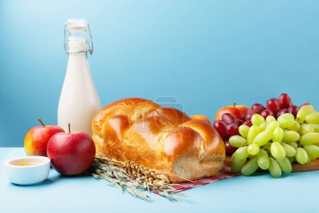 Foto de Tarjeta festiva de Shavuot feliz. Concepto de fiesta religiosa judía. Productos lácteos, manzanas frescas, uvas, queso, pan, leche, requesón, trigo y miel sobre fondo azul, espacio para copiar. - Imagen libre de derechos