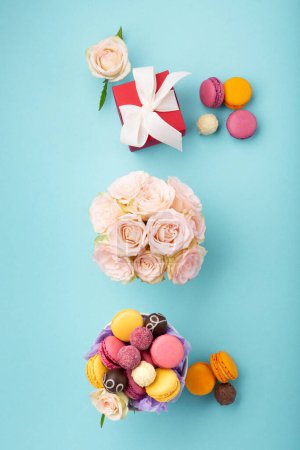 Photo for Sweet cookies macaroons, red gift box, rose flowers and chocolate candies on blue background. Spring presents concept for Mothers day, womans day, birthday. Flat lay, empty space for text or message - Royalty Free Image