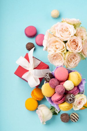 Photo for Sweet cookies macaroons, red gift box, rose flowers and chocolate candies on blue background. Spring presents concept for Mothers day, womans day, birthday. Top view, copy space - Royalty Free Image