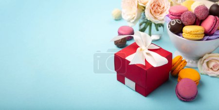 Photo for Sweet cookies macaroons, red gift box, rose flowers and chocolate candies on blue background. Spring presents concept for Mothers day, womans day, birthday, empty space for text or message - Royalty Free Image