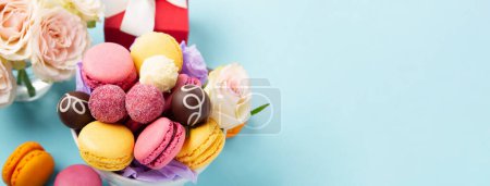 Photo for Sweet cookies macaroons, red gift box, rose flowers and chocolate candies on blue background. Spring presents concept for Mothers day, womans day, birthday. Banner, empty space for text. - Royalty Free Image
