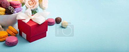 Photo for Sweet cookies macaroons, red gift box, rose flowers and chocolate candies on blue background. Spring presents concept for Mothers day, womans day, birthday, empty space for text or message - Royalty Free Image