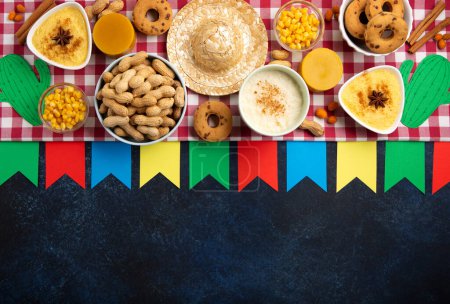 Festa Junina Sommerfest Karnevalskonzept. Brasilianischer Strohhut, Maiskuchen, Erdnüsse und bunte Fahnen auf blauem Holzgrund, Draufsicht. Design für Grußkarte, Einladung oder Feiertagsbanner