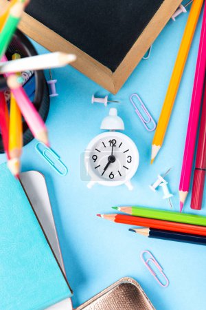 Photo for Back to school, Exam, Education concept on blue background. School scattered stationery, notepads, pens, pencils, paper clips and white alarm clock on blue table. Top view, copy space. - Royalty Free Image
