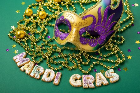 Mardi Gras King Cake cookies, masquerade festival carnival mask, gold beads and golden, green, purple confetti on green background. Holiday party invitation, greeting card concept.