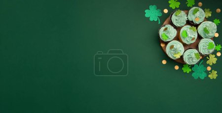 Foto de Día de San Patricio festivo cupcakes de chocolate de vainilla con glaseado verde y decoraciones de trébol brillante sobre fondo de papel verde. Concepto de postre navideño irlandés. Vista superior, espacio de copia. - Imagen libre de derechos