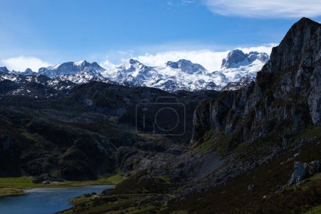 A stunning view of the Lakes of Covadonga in the foreground, with the towering peaks of the Picos de Europa mountain range in the background. The calm water reflects the rugged beauty of the mountains under a clear sky.