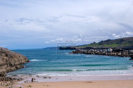A sandy beach stretches alongside the ocean under a cloudy sky, with waves gently rolling onto the shore. In the distance, mountains of Llanes, Asturias, can be seen