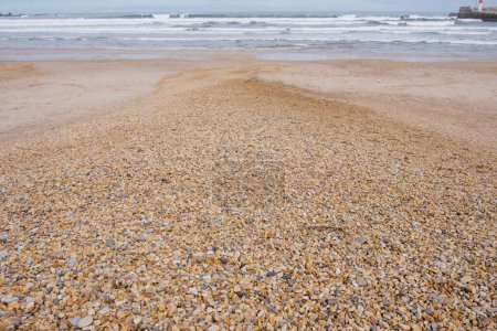 Ein Sandstrand mit nassen Kieselsteinen am Ufer, in der Ferne ein Boot auf dem ruhigen Wasser des Ozeans.