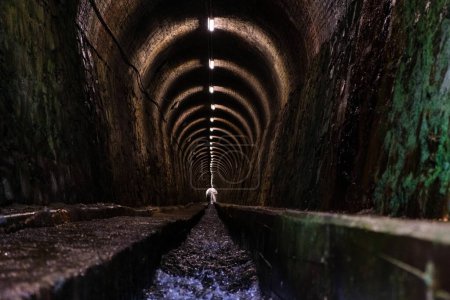 largo túnel, iluminado por luces artificiales, rodeado de paredes a cada lado, canal de agua