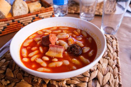 Auf einem Holztisch steht eine Schüssel gefüllt mit Fabada Asturiana, einer traditionellen spanischen Bohnen- und Fleischsuppe. Die Suppe ist herzhaft und würzig, enthält zarte Fleischstücke und cremige Bohnen.