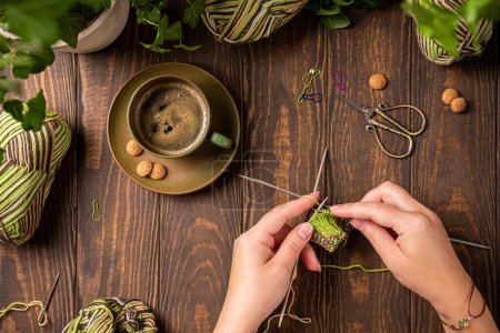 Photo for Flat lay with homemade knitting green socks in progress with female hands. Composition on wooden table with coffee cup and house plants, above, top view. - Royalty Free Image