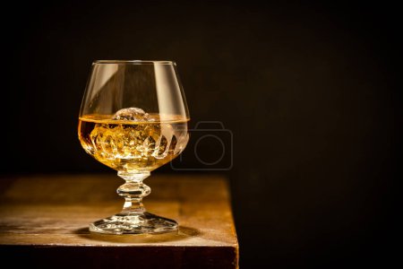 Whiskey with ice or brandy in glass on rustic wooden table over black background with copy space