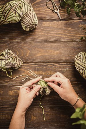 Photo for Flat lay with female hands knitting green socks in progress. Cozy homely atmosphere with wooden table and house plants, copy space, overhead shot. - Royalty Free Image