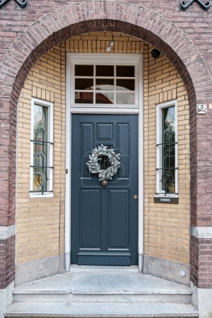 Photo for Elegance and classic design of dark, wooden front door adorned with a decorative wreath, set within arched entrance of a brick building. The door is complemented by two small windows on either side - Royalty Free Image
