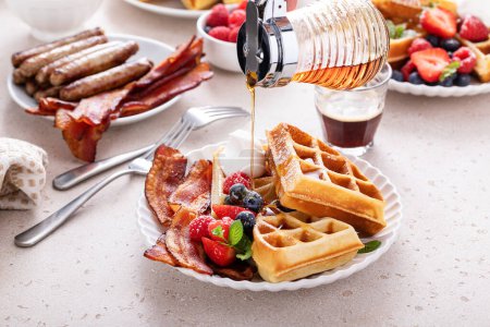 Photo for Breakfast table with waffles, croissants, sausage and bacon served maple syrup pouring over - Royalty Free Image