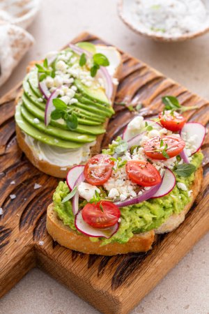 Avocado toasts on a wooden board with cream cheese, radishes and feta topped with red onion and freshly ground pepper