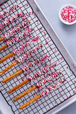 Chocolate dipped pretzel rods with dark and white chocolate and pink heart sprinkles on a cooling rack