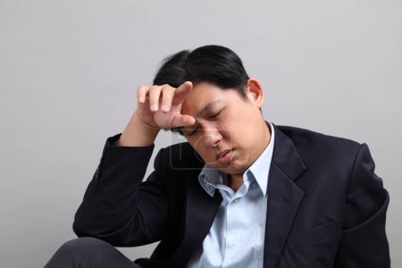 El empresario asiático con formal vestido mientras se sienta con gesto de agotado sobre el fondo gris.