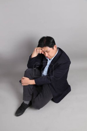 Der asiatische Geschäftsmann mit formalen gekleidet, während er mit Geste der Erschöpfung auf dem grauen Hintergrund sitzt.