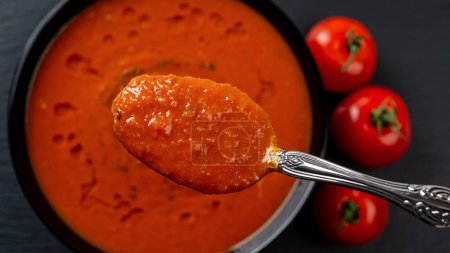 Photo for Spoon of hot tomato soup over bowl closeup. Eating vegetable dish of pureed roasted tomatoes, garlic and basil. Healthy vegetarian dish. Mediterranean cuisine. Focus on spoon. Top view. - Royalty Free Image