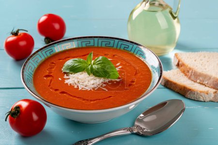 Photo for Tomato soup bowl and spoon over turquoise wooden table. Homemade hot soup of roasted tomatoes with parmesan cheese and green basil. Mediterranean cuisine. Healthy vegetarian dish. Front view. - Royalty Free Image
