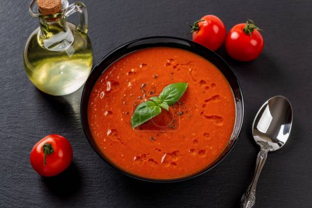 Photo for Hot tomato soup and spoon over black background. Portion of freshly made soup puree of roasted tomatoes, garlic and basil in a black bowl. Healthy vegan and vegetarian dish. Top view. - Royalty Free Image