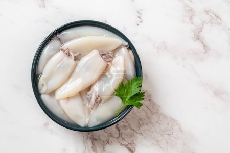 Raw squid fillet in a bowl over marble background. Small calamary tubes prepared for cooking. Fresh squid molluscs ingredient for low calorie healthy diet. Seafood concept. Top view.