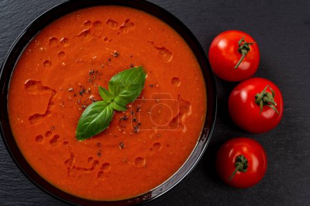 Photo for Tomato soup puree in a black bowl closeup. Freshly made hot vegetable dish of pureed tomatoes, garlic and basil. Mediterranean cuisine. Healthy vegan and vegetarian recipe. Top view. - Royalty Free Image