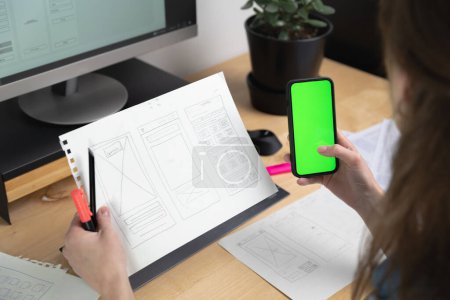 Téléchargez les photos : Sketch of wireframe for website. Women designer working on a mobile application, holding in hand phone with green screen - en image libre de droit
