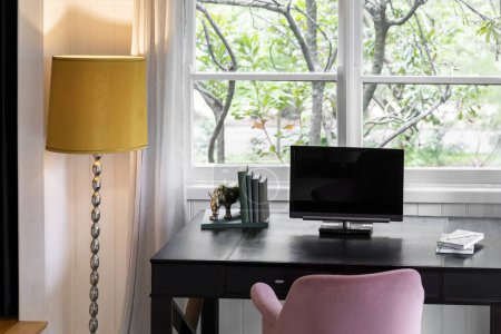 Téléchargez les photos : Bureau à domicile moderne doté d'un ordinateur, de livres et d'une lampe jaune électrique, le tout face à une fenêtre pour un espace de travail élégant et fonctionnel. - en image libre de droit