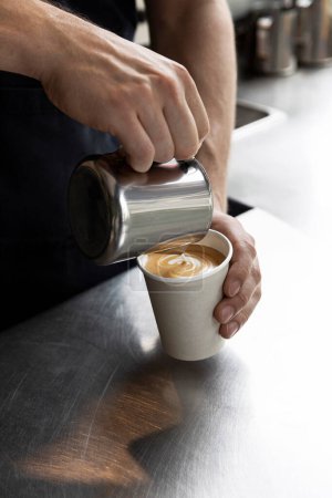Téléchargez les photos : Gros plan des mains d'un barista versant du lait dans une tasse à café, créant un latte art dans une cuisine commerciale. - en image libre de droit