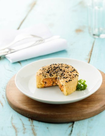 Ein Teller mit herzhaftem Hähnchenkuchen, mit knuspriger goldbrauner Kruste und schwarzem und weißem Sesam darauf, fertig zum Essen. Auf einem blauen Holztisch.