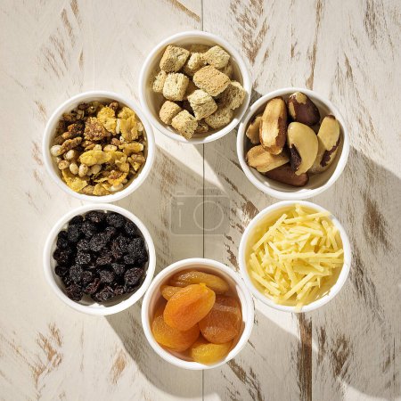 Téléchargez les photos : Six ramequins aux céréales, croûtons, noix du Brésil, parmesan, abricot turc et raisins secs, sur une table en bois blanc - en image libre de droit