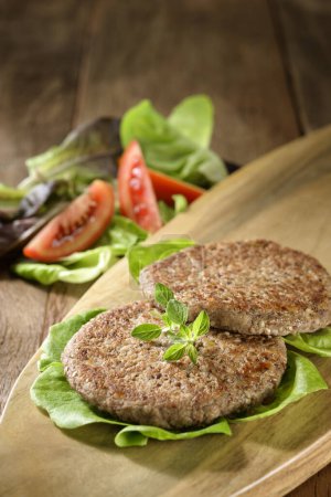 Photo for Two delicious vegetarian or meat burger served on a cutting board with lettuce salad and tomato aside, on a wooden table - Royalty Free Image