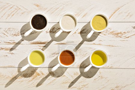 Seis botes de salsa o ramekins con diferentes tipos de salsas, encima de una mesa rústica de madera blanca.