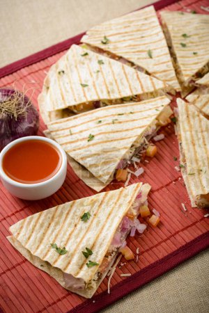 Téléchargez les photos : Quesadilla épicée à base de tortilla au fromage, tomate et oignons, avec une sauce aux épices à côté pour tremper. Sur une table rouge tapis. - en image libre de droit