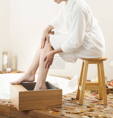Foto de Mujer empapándose los pies, un baño de pies terapéutico que puede ser muy relajante, en un cuenco de madera junto a la bañera. - Imagen libre de derechos