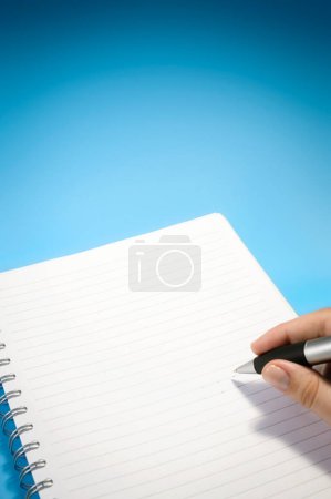 Foto de Mano sosteniendo un bolígrafo, a punto de escribir en un papel con líneas de un cuaderno. El fondo es azul con un gradiente. - Imagen libre de derechos