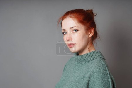 mujer joven con moño de pelo rojo y jersey de cuello de rollo verde contra fondo gris con espacio para copiar