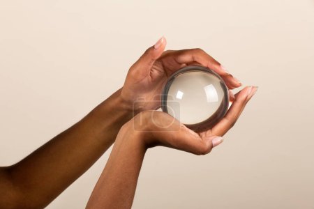 Foto de Manos de mujer negra sosteniendo bola de cristal transparente con reflejo de luz sobre fondo beige - Imagen libre de derechos