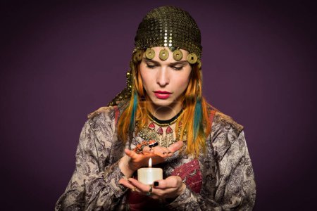 Photo for Serious female fortune teller in gypsy outfit and headwear putting hand over light candle for predicting future against dark background - Royalty Free Image