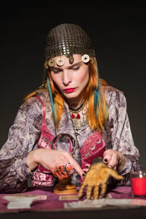 Photo for Serious redhead female fortune teller in gypsy outfit and headwear showing prediction of future on artificial palm against dark background - Royalty Free Image