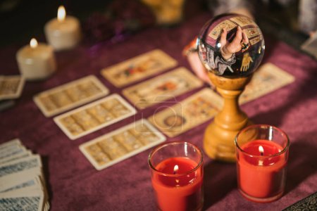 Selektiver Fokus der Kristallkugel reflektiert Erntewahrsager, der die Zukunft vorhersagt, mit Tarotkarten in der Nähe brennender Kerzen auf dem Tisch vor verschwommenem Hintergrund