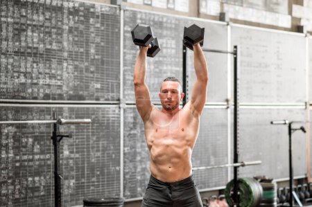 Photo for Shirtless muscular male athlete lifting dumbbells over head while performing devil press exercise at gym - Royalty Free Image
