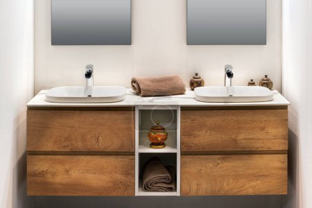 Interieur des modernen minimalistischen Badezimmers mit doppelten weißen Waschbecken Wasserhähne Handtuch auf hölzernen Schrankablagen mit Räucherstäbchen Vase platziert, während Spiegel an der weißen Wand befestigt